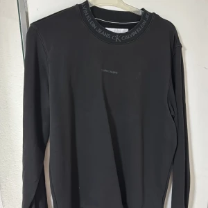 Svart sweatshirt från Calvin Klein Jeans - Svart långärmad sweatshirt från Calvin Klein Jeans med logotyp tryckt på bröstet och runt halsringningen. Tröjan har en klassisk rund hals och är gjord i mjuk bomull, perfekt för en chill och stilren look. Passformen är normal pch storlek M 