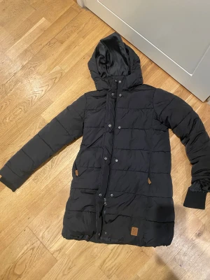 Svart lång pufferjacka från x-trail - Säljer en svart pufferjacka från xtrail med huva och långa ärmar. Jackan har quiltad design, dragkedja och knappar framtill samt två fickor med snygga detaljer. Perfekt för kalla dagar och har en stilren look som funkar till det mesta.
