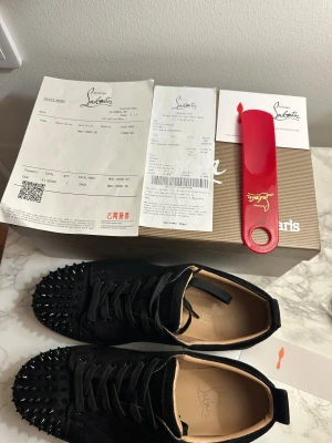 Christian Louboutin svarta sneakers med nitar - Svarta sneakers från Christian Louboutin i mocka med rund tå och platt sula. Tåpartiet är täckt av svarta nitar för en edgy look. Skorna har snörning och den klassiska röda sulan med Louboutin-logga. Perfekt för dig som vill sticka ut med lyxig streetstyle. OPPS använda storlek 43 pris kan diskuteras