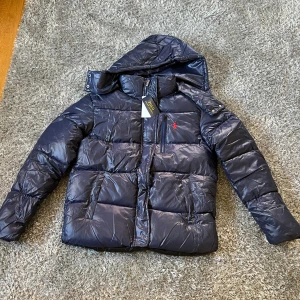 Mörkblå pufferjacka från Polo Ralph Lauren - Snygg mörkblå pufferjacka från Polo Ralph Lauren med huva och röd broderad logga på bröstet. Jackan har dragkedja framtill och quiltad design som ger en cool look. Perfekt för kalla dagar när du vill vara både varm och stilren. Pris kan diskuteras. fickan är ihopsydd därför säljer den för lågt  