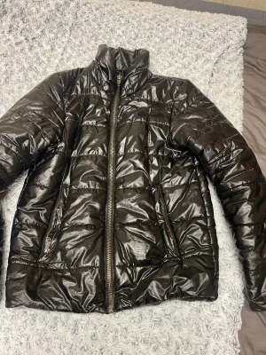 Svart glansig pufferjacka - Säljer en svart glansig pufferjacka med hög krage och dragkedja framtill. Jackan har två fickor med dragkedja och quiltad design. Perfekt för kyligare dagar och ger en cool look till din outfit.