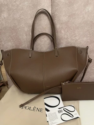 Brun axelväska från Polène i skinn - Snygg brun axelväska från Polène med stilren design och vita kontrastsömmar. Väskan är tillverkad i strukturerat skinn och har både korta och långa handtag samt en matchande plånbok. Perfekt storlek för allt du behöver och riktigt chic form.