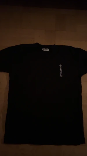 Svart Moncler t-shirt med logga - Svart t-shirt från Moncler med vit logotyp tryckt vertikalt på bröstet. Klassisk rund hals och korta ärmar. Materialet är mjuk bomull och på ena ärmen finns en diskret Moncler-patch. Perfekt för dig som gillar stilrena designer.