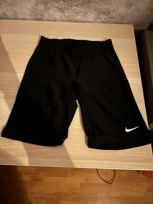 Svarta träningsshorts från Nike - Svarta shorts från Nike med klassisk vit swoosh-logga på benet. Modellen har elastisk midja och är tillverkad i mjuk polyester som andas. Perfekta för sport eller chill, med enkel och stilren design utan mönster eller extra detaljer.