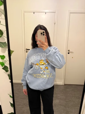 Blå Garfield sweatshirt med tryck - Säljer en ljusblå sweatshirt med Garfield-tryck där han spelar basket. Tröjan har rund hals, långa ärmar och en loose passform. Mjuk insida och ribbade muddar vid ärmslut och nederkant. .