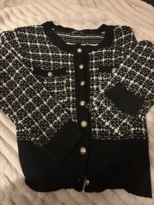 Svartvit rutig kofta från SHEIN - Snygg svartvit rutig kofta från SHEIN med guldfärgade knappar framtill och på fickorna. Koftan har rund hals, långa ärmar och breda ribbade muddar vid ärmslut och nederkant. Materialet är stickat och ger en mjuk och skön känsla. Denna tröjan kommer ej till användning då jag helt änkelt glömde bort den och sen slutade använda. 