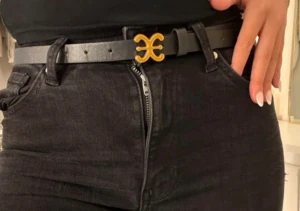 Svart skärp med guldspänne - Snyggt svart bälte med ett unikt guldspänne i en cool form. Bältet är smalt och passar perfekt till jeans eller byxor. Materialet ser ut som skinn och spännet ger en trendig touch till din outfit.