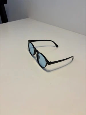 Chimi liknande solglasögon  - Snygga svarta glasögon med rund form och blåtonade linser. Bågarna är tunna och stilrena, perfekta för dig som gillar en clean och modern look. Passar dig som vill sticka ut med färgade linser och klassisk design. Helt nya.
