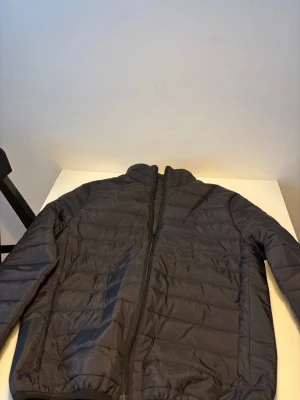 Svart quiltad pufferjacka herr - Svart pufferjacka med quiltad design och hög krage. Jackan har hel dragkedja framtill och är tillverkad i syntetmaterial. Perfekt för dig som vill ha en clean och enkel look under kyliga dagar. Helt ny.