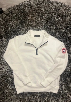 Vit half zip sweatshirt Canada Goose - Vit sweatshirt från Canada Goose med half zip och klassisk logga på vänster ärm. Tröjan har lång ärm, ribbade muddar och är gjord i mjuk bomull. Perfekt för dig som gillar clean och stilren look med premiumkänsla.