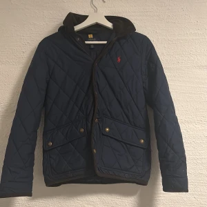 Mörkblå quiltad jacka från Polo Ralph Lauren - Snygg mörkblå quiltad jacka från Polo Ralph Lauren med huva och bruna detaljer. Jackan har två stora fickor med knappar, dragkedja och tryckknappar framtill samt röd logga på bröstet. Perfekt för höst och vinter när du vill vara både varm och stilren.luvan går att ta av och på. Pris går att diskuteras. 14-16 ålder