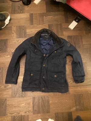 Mörkblå vadderad fieldjacka från Marc O'Polo - Riktigt snygg mörkblå vadderad field jacka från Marc O'Polo med quiltat foder och flera stora fickor framtill. Jackan har en krage i brun mocka-look och stängs med både knappar och dragkedja. Perfekt för vintern. Vid frågor eller funderingar, fråga på! 