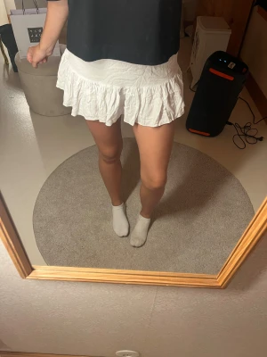 Vit volangkjol  - Supersöt vit kortkjol med bred smock i midjan och dubbla volanger nertill. Kjolen är luftig och har en lätt rynkad struktur som ger en avslappnad och trendig look. Perfekt att styla med en enkel topp eller hoodie.