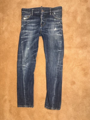  Dsquared2 Jeans - Snygga blå jeans från Dsquared2 med coola slitningar och blekta detaljer. Klassisk femficksmodell med Dsquared2-logga vid fickan och brunt läderpatch bak. Jeansen har raka ben och normal passform, tillverkade i slitstarkt denimtyg. Skick 8-10. Nypris 4499kr Mitt pris 299kr. Storlek 42