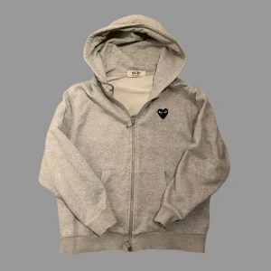 Grå hoodie från Comme des Garçons Play - Grå hoodie med dragkedja från Comme des Garçons Play. Tröjan har huva och ett svart hjärta med ögon på bröstet samt ett stort hjärta med ögon tryckt på ryggen. Tillverkad i mjuk bomull, perfekt för chill dagar. Klassisk passform och ribbade muddar.