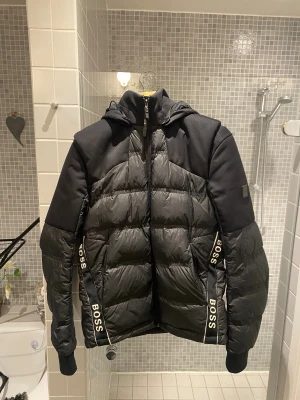 Svart pufferjacka från BOSS med huva - Svart pufferjacka från BOSS med avtagbara ärmar så du kan bära den som väst. Jackan har huva, dragkedja framtill och BOSS-logga på ärmen och längs sidorna. Materialmix med polyester och syntet, perfekt för kalla dagar. Snygg och sportig look med modern passform.  Priset går att diskutera. 
