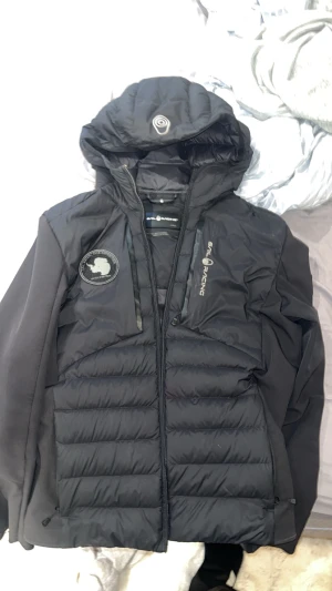 Sail Racing jacka - Säljer en svart dunjacka från Sail Racing med huva och snygga loggor på bröstet och ärmen. Jackan har quiltad design, dragkedja framtill och två sidofickor. Perfekt för kalla dagar och riktigt clean look. Lite trasig vid armen och dragkedjan, dragkedjan har gåttt av men den medföljer med jackan, jackan går ej knäppa men den går att laga