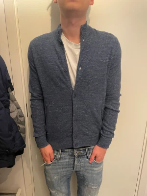 Stickad cardigan  - Snygg blåmelerad kofta från Ollygan i storlek S. Koftan har en ståkrage och stängs med knappar framtill. Materialet är mjukt och stickat, perfekt för att ge en chill och stilren vibe. Passar dig som gillar enkel och clean look.