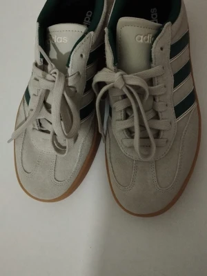 Adidas Samba sneakers ljusgrå/grön - Säljer ett par Adidas Samba sneakers i ljusgrå mocka med gröna och vita detaljer. Skorna har klassiska tre ränder på sidan, snörning och en gummisula i brunt. Insidan är fodrad med mörkgrönt tyg och tån är rundad. Perfekt för dig som gillar retrostil och streetwear.