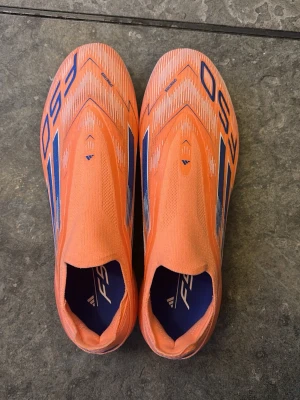 Adidas F50 orange fotbollsskor - Säljer ett par Adidas F50 fotbollsskor i en riktigt snygg orange färg med blå detaljer och grafiskt mönster. Skorna har en strömlinjeformad design utan snörning och är tillverkade i syntetmaterial med Sprintweb-teknologi för extra lätthet och snabbhet på planen.