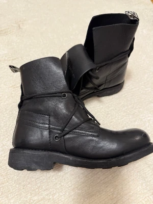 Svarta kängor från Bikkembergs - Säljer ett par svarta kängor från Bikkembergs i skinn med grov sula där loggan är präglad undertill. Kängorna har snörning och hög skaft med tydlig Bikkembergs-text på plösen. Perfekt för dig som gillar robust stil och snygga detaljer.