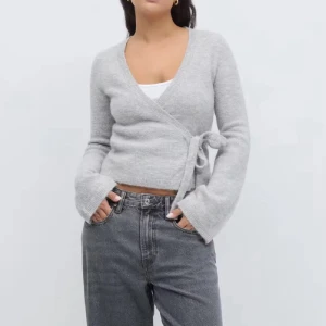 Grå omlottkofta med knyt - Säljer en mjuk och mysig grå omlottkofta med knyt i sidan. Koftan har v-ringad hals och långa ärmar med lätt utsvängda muddar. Perfekt att styla med jeans eller kjol för en chill look.