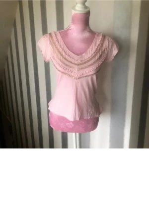 Ljusrosa blus med volang och guld - Supersöt ljusrosa blus med korta ärmar och v-ringad hals. Dekorerad med glittriga guldiga band och volanger både fram och bak. Blusen har en feminin passform och är gjord i mjukt bomullsmaterial. Perfekt för dig som gillar romantiska detaljer och pastellfärger.