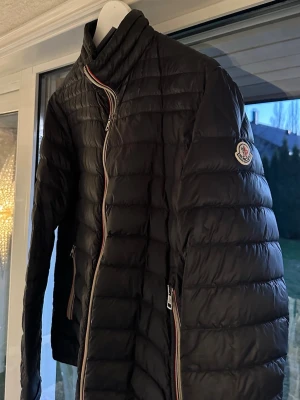 Svart Moncler pufferjacka - Säljer en svart pufferjacka från Moncler med klassisk quiltad design och snygga detaljer i form av röd, vit och blå randig dragkedja. Jackan har hög krage, två fickor med dragkedja och Moncler-logga på ärmen. Perfekt för kyliga dagar och riktigt stilren.