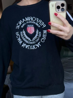 Mörkblå stickad tröja Performance - Snygg mörkblå stickad tröja från Performance med broderat emblem och text på bröstet. Tröjan har rund hals, ribbade muddar och vita detaljer längs ärmarna. Perfekt för dig som gillar preppy stil och vill ha något som sticker ut.