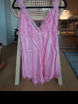 Rosa linnetopp med spetsdetaljer - Säljer en ljusrosa linnetopp med v-ringad hals och breda axelband. Toppen har fina spetsdetaljer längs halsringningen och nederkanten som ger en romantisk vibe. Materialet är mjukt och glansigt, troligen polyester eller satin. Perfekt för sommarens varma dagar.