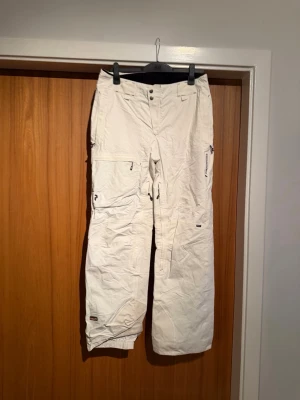 Peak performance Vita täckbyxor i Gore-Tex - Säljer ett par vita täckbyxor med flera praktiska fickor och dragkedjor. Byxorna är tillverkade i slitstarkt Gore-Tex-material som står emot väta och vind. De har normal passform, elastiska benslut och detaljerade sömmar. Perfekt för vinterns äventyr.