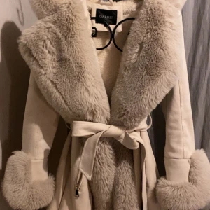 Beige pälsjacka från Garoff med bälte - Superfin beige pälsjacka från Garoff med stora fluffiga slag, pälsdetaljer på ärmsluten samt luva och ett matchande bälte i midjan. Jackan har dragkedjor vid fickorna och är perfekt för kalla dagar när du vill vara både varm och snygg.