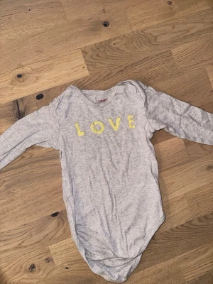Grå body med LOVE-tryck från Livly Livly grå body, storlek 62/68 - Grå baby body med långa ärmar och texten "LOVE" tryckt på framsidan. Säljer en ljusgrå body från Livly med lång ärm och tryckta gula bokstäver som bildar ordet LOVE på bröstet. Bodyn är tillverkad i mjuk bomull och har praktiska tryckknappar i grenen för enkel på- och avklädning.