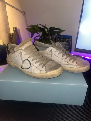Philippe Model vita sneakers - Säljer ett par Philippe Model sneakers i vitt med grå detaljer och svart häl. Skorna har snörning, rund tå och platt sula. Materialet är skinn och mocka, med en ikonisk sköld på sidan. Perfekta för dig som gillar stilrena och trendiga sneakers.