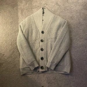 Grå stickad kofta med knappar - Säljer en grå stickad kofta med bred krage och stora svarta knappar framtill. Koftan har långärmad design och ribbade muddar vid ärmslut och nederkant. Perfekt för lager-på-lager och chill dagar.