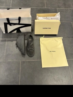 Grå sneakers från Axel Arigato - Säljer ett par grå sneakers från Axel Arigato med snygga detaljer i mocka på tå och häl. Skorna har snörning, platt sula och rund tå. Märket syns i guldtext på sidan och bak. Kommer med originalkartong och dustbag.                                 Nypris 4000kr