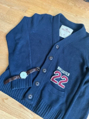 Mörkblå kofta från Hollister med tryck - Snygg mörkblå kofta från Hollister i storlek M men som sitter mer som S. Stickad i mjukt material med ribbade muddar och knappar framtill. På bröstet finns ett broderat Hollister-tryck och siffran 22 i rött och vitt. Perfekt för lager-på-lager och chill stil.