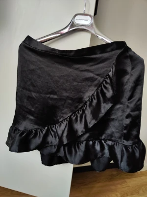 Svart omlottkjol med volang - Snygg svart kort omlottkjol i glansigt satinliknande material med bred volang längs nederkanten. Kjolen har en feminin och trendig look som passar perfekt till sneakers eller boots. Elastisk midja för skön passform.