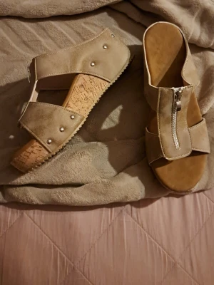 Beige sandaler med dragkedja och nitar - Säljer ett par snygga beige sandaler med kilklack i kork och öppen tå. Ovandelen är i mockaimitation med silverfärgade nitar och dragkedja framtill. Perfekta för sommaren och ger en cool touch till din outfit. De är en 41 men är mer som en 40