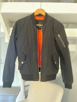 Svart bomberjacka från ONLY - Svart bomberjacka med orange foder från ONLY. Jackan har klassisk ribbad mudd vid ärmslut och midja, dragkedja framtill och fickor med tryckknappar. På ena ärmen finns en zip-detalj. Materialet är polyester och jackan är långärmad.