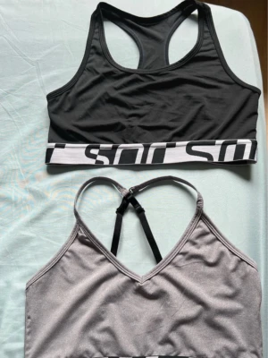 Två sport-bh från SOC, svart & grå - Säljer två sport-bh från SOC i storlek M. En är svart med bred vit och svart logotypkant, den andra är ljusgrå med samma kant och svarta justerbara axelband. Båda är gjorda i mjuk polyester och elastan, perfekta för träning.