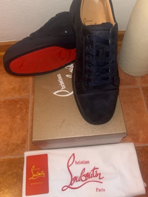 Svarta sneakers från Christian Louboutin - Säljer ett par svarta sneakers från Christian Louboutin med klassisk röd sula och rund tå. Skorna har snörning och är tillverkade i mocka med snygga detaljer. Kommer med originalkartong, dustbag och äkthetskort. Perfekt för dig som gillar lyxiga och stilrena sneakers.