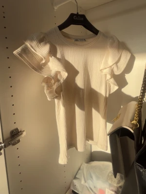 Ribbad vit topp med volang från Zara - Säljer en ribbad beige topp från Zara med korta ärmar och stora volangdetaljer på axlarna. Toppen har rund halsringning och är gjord i ett mjukt material som känns skönt mot huden. Perfekt för dig som gillar en feminin och trendig stil.