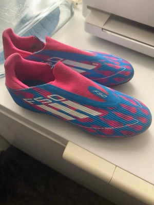 Adidas F50 blå/rosa fotbollsskor - Säljer ett par Adidas F50 fotbollsskor i en riktigt fet färgkombo med blå och rosa mönster och vita detaljer. Skorna har platt sula med dobbar för gräsplan och är tillverkade i syntetmaterial. Snygg, strömlinjeformad design utan snörning för max speed på planen.