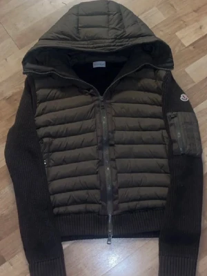 Moncler cardigan  - Säljer en mörkbrun pufferjacka från Moncler med stickade ärmar och huva. Jackan har quiltad front, dragkedja och en ficka med logga på ärmen. Perfekt för kyliga dagar och har en snygg, urban vibe.