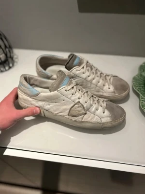 Philippe Model vita sneakers med blå detalj - Säljer ett par vita Philippe Model sneakers med ljusblå detalj vid hälen och tungan. Skorna har rund tå, snörning och platt sula med tydlig PHILIPPE-logga undertill. Materialet är skinn och mocka, vilket ger en cool och avslappnad vibe.