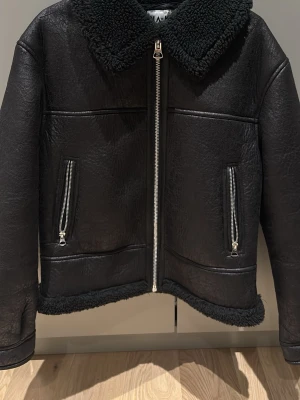 Svart teddyjacka från NA-KD - Säljer en svart teddyjacka från NA-KD i storlek 36. Jackan har grov krage och foder i teddy, två fickor med dragkedja och detaljer i skinnimitation. Perfekt för kalla dagar med sin mysiga och coola look. Dragkedja framtill och snyggt avslut vid nederkant.