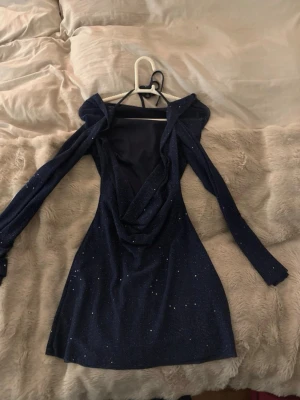 Glittrig marinblå kortklänning - Säljer en snygg marinblå kortklänning med glittriga detaljer. Klänningen har långärmade ärmar och en draperad omlottfront som ger en cool look. Materialet är mjukt och stretchigt, perfekt för att sticka ut. Passformen är tight och klänningen har en elegant v-ringning.