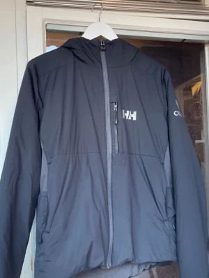 Svart vindjacka från Helly Hansen - Snygg svart vindjacka från Helly Hansen med huva och hel dragkedja framtill. Jackan har en bröstficka med dragkedja och diskret logga på bröstet. Perfekt för blåsiga dagar och outdoor-aktiviteter. Lätt och smidig design med lång ärm.