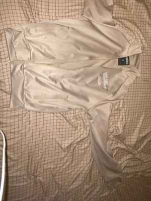 Beige ziptröja från Hoodrich - Säljer en beige ziptröja från Hoodrich med vit logga på bröstet och vita ränder längs ärmarna. Tröjan har lång ärm, ribbade muddar och dragkedja framtill. Materialet är mjukt och har en lätt glansig finish, perfekt för chill eller träning.